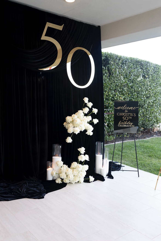 A 50th birthday party backdrop setup featuring a large black draped curtain with a metallic gold '50' and white floral arrangements on a white floor, with a welcome sign on an easel nearby.